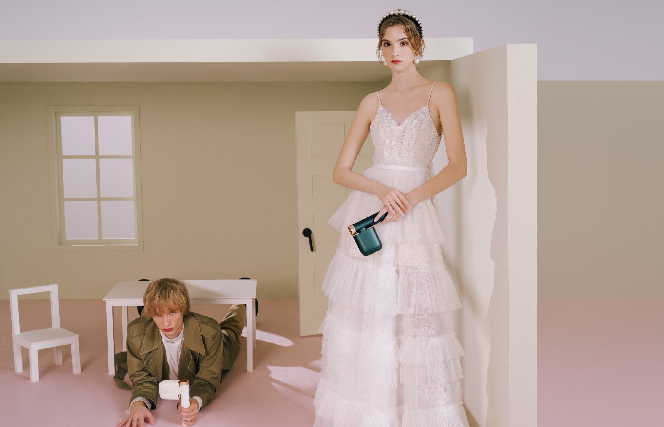 Fashion shoot depicting a model in a tiered wedding gown standing next to a man under a table, both showcasing at home IPL hair removal gadgets.