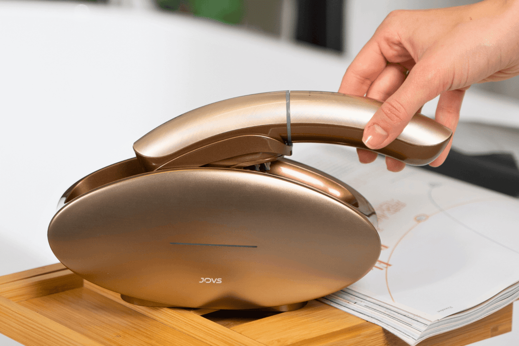 Hand lifting JOVS Slimax beauty device in gold, set on a wooden tray over an open book