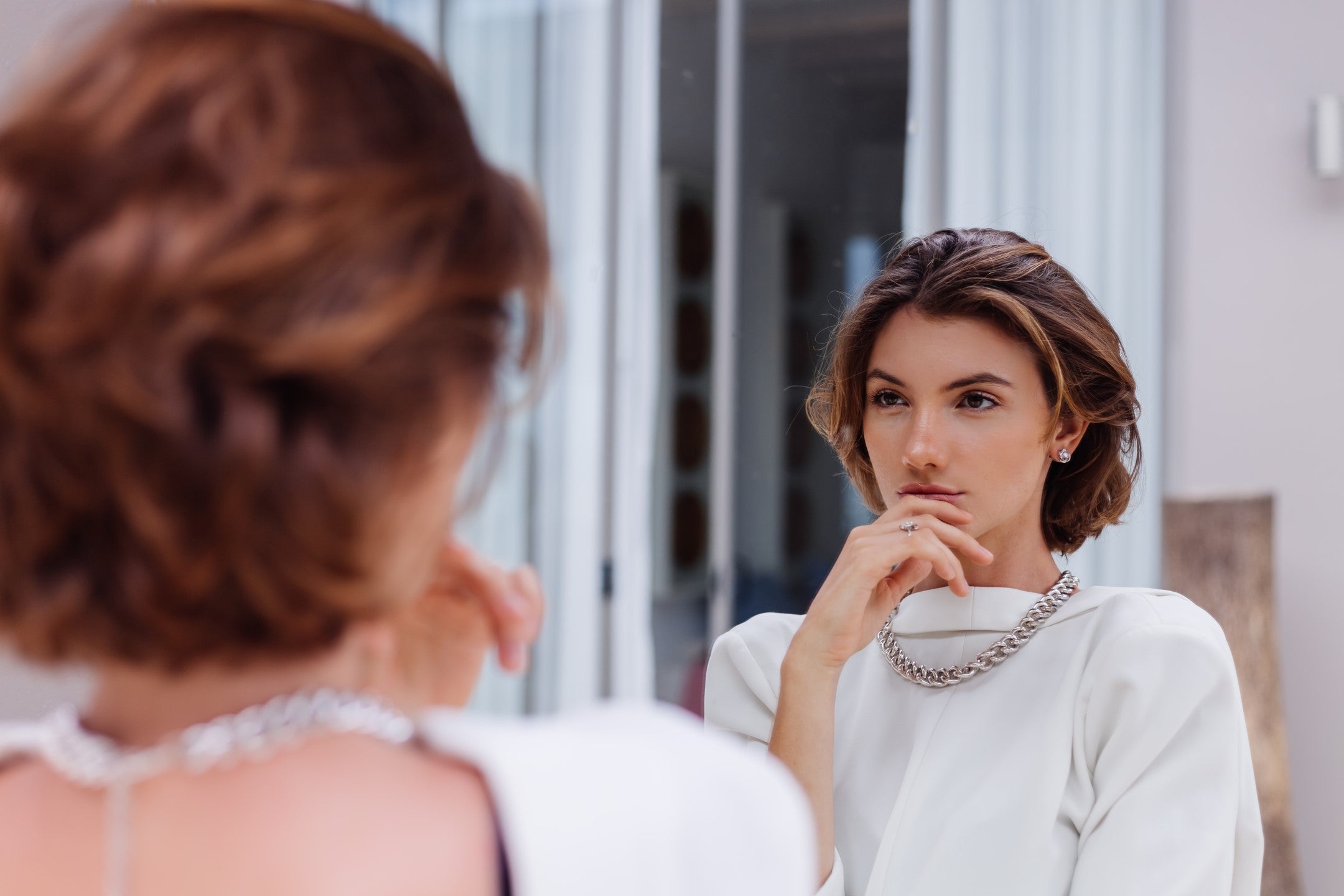 Woman in white blazer contemplating how to stop facial hair growth naturally while looking in mirror at luxury villa.