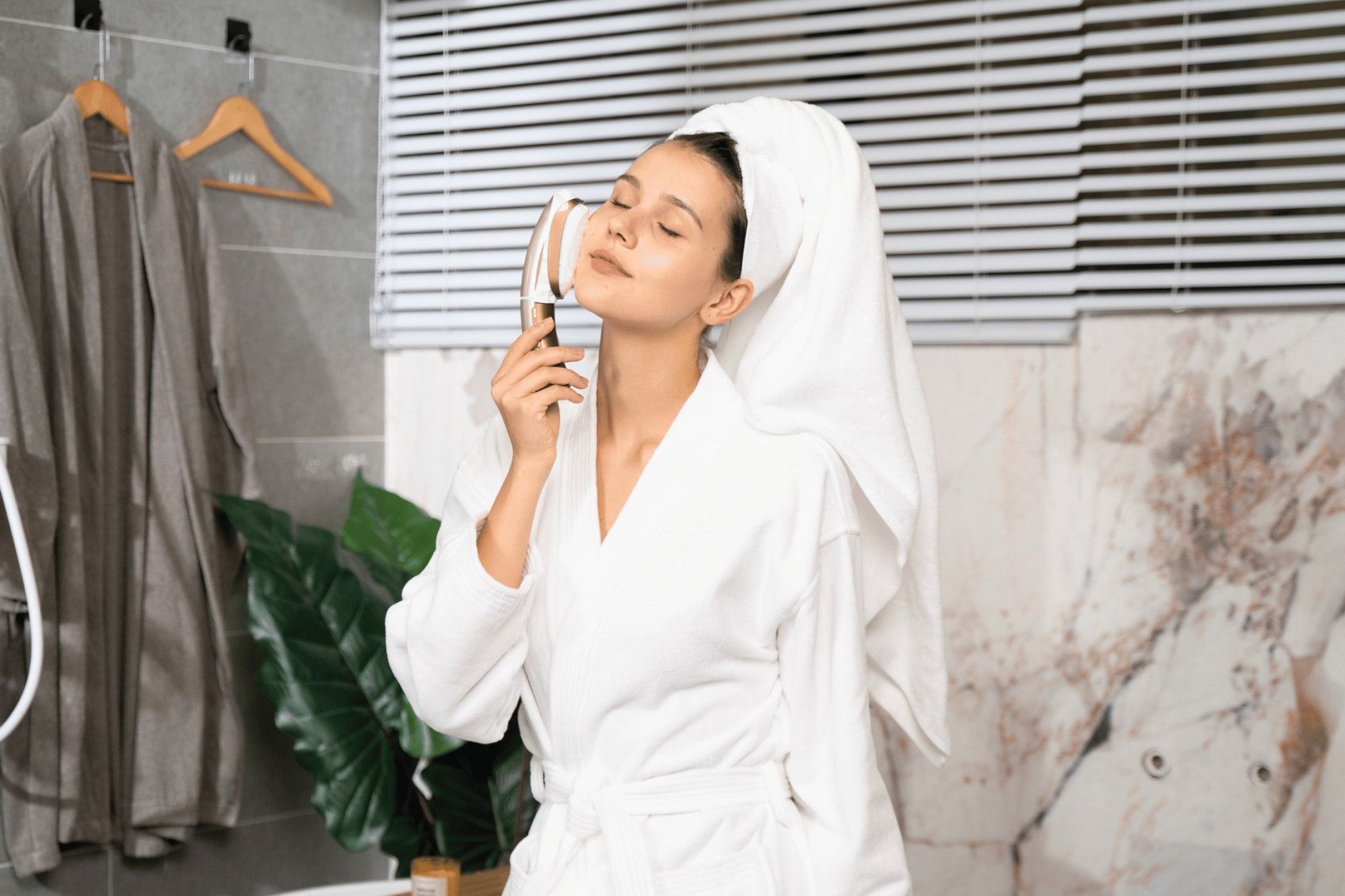 A relaxed woman in a bathrobe and towel is performing a microcurrent facial massage with a handheld beauty device.