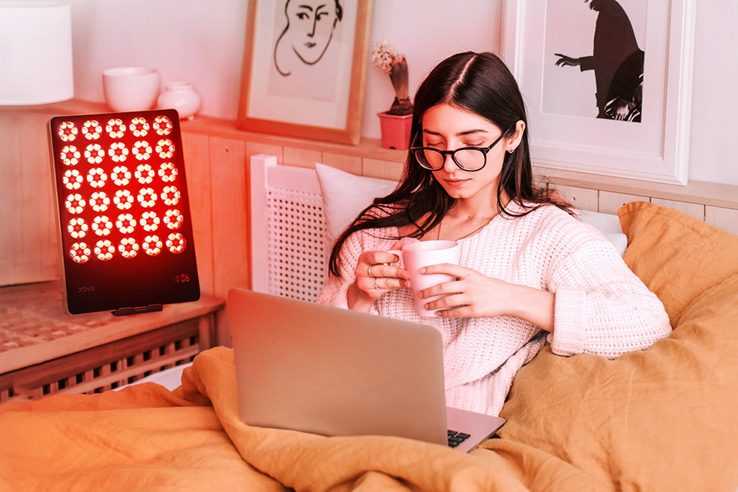 Woman in glasses sitting on bed with laptop and cup, red light therapy device on bedside table.